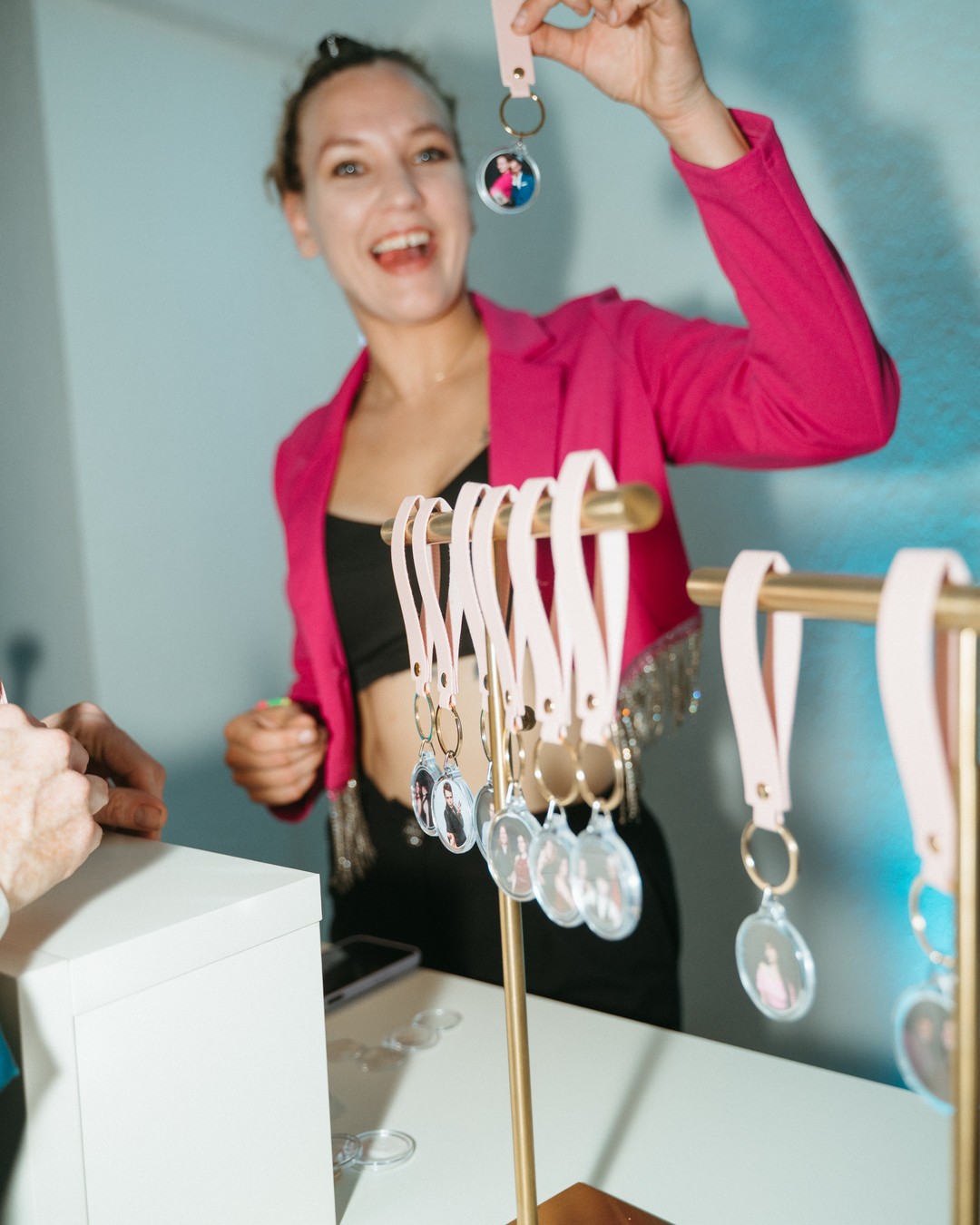 Smiling woman in a pink blazer holding up a finished photo keychain above a rack of blush straps